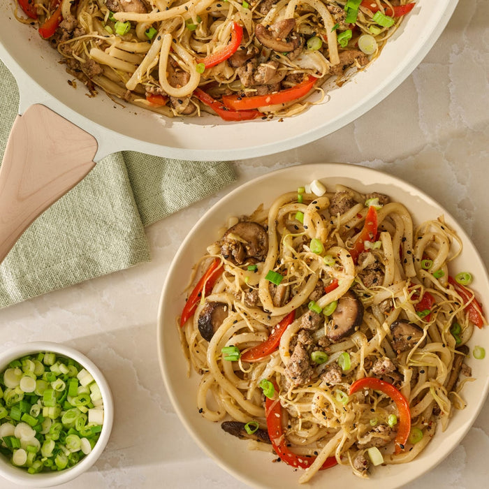 A plated serving of udon noodle stir-fry, made with Mccormick Sesame Ginger Seasoning, 2.5 Oz, sits next to the pan and a bowl of chopped green onions.