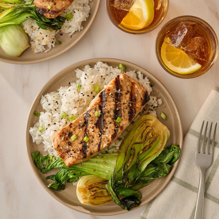 An overhead shot shows a plate of grilled salmon seasoned with Mccormick Sesame Ginger Seasoning, 2.5 Oz, served on a bed of white rice with grilled bok choy, next to a glass of iced tea.