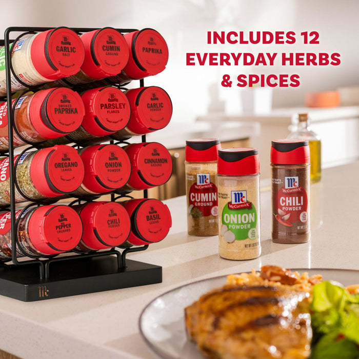 A Mccormick Spice Rack Red Cap Black Metal 12 Ct, holding a variety of McCormick herbs and spices such as garlic salt, paprika, thyme, oregano, and cinnamon, is displayed on a kitchen counter alongside three individual McCormick jars of cumin ground, onion powder, and chili powder, with a prepared meal in the foreground.