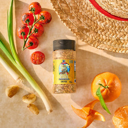 A bottle of Mccormick, 10.49 Oz all-purpose seasoning, featuring a limited edition One Piece label, is arranged in a flat lay with charred tomatoes, scallions, an orange, and a straw hat.