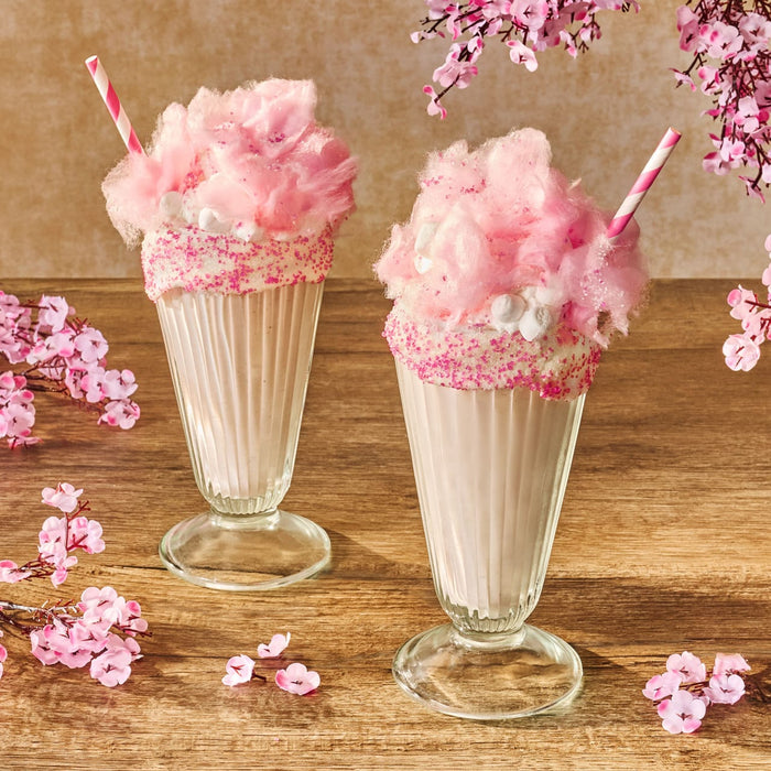 Two festive pink milkshakes in tall glasses are topped with cotton candy and decorated with pink sprinkles from a package of Mccormick, 10.49 Oz, all set on a wooden table with cherry blossoms.