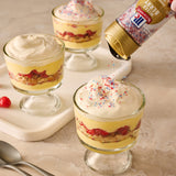 Dessert glasses with layers of yellow pudding, red fruit, and whipped cream being topped with colorful sprinkles.