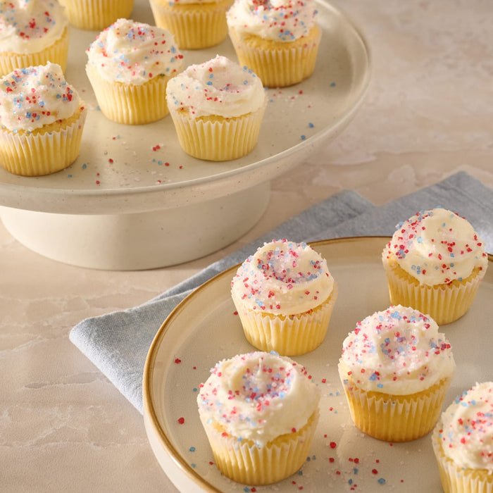 A collection of mini vanilla cupcakes with white frosting are beautifully decorated with a sprinkle of Mc Mixed Berry Fin Sgr 72/6/3.15 Oz, arranged on a cake stand and plate.
