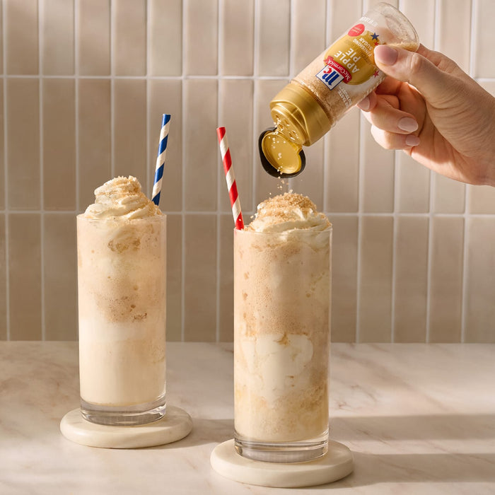 A hand sprinkles Mccormick Apple Pie Finishing Sugar, 3.06 Oz, over a tall glass of an apple pie milkshake topped with whipped cream, with a second milkshake visible nearby.