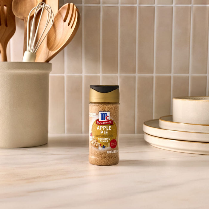 A bottle of Mccormick Apple Pie Finishing Sugar, 3.06 Oz, is displayed on a kitchen countertop next to a utensil holder and a stack of plates, creating a warm baking scene.