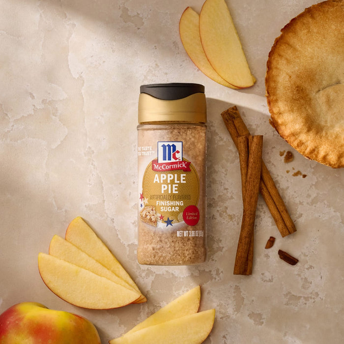 A bottle of Mccormick Apple Pie Finishing Sugar, 3.06 Oz, is arranged on a light-colored surface with fresh apple slices, cinnamon sticks, and a golden-brown apple pie.