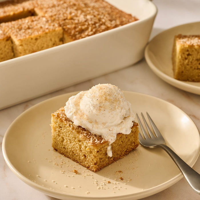 A square slice of light brown cake sits on a beige plate, topped with a scoop of cream and a sprinkle of Mccormick Apple Pie Finishing Sugar, 3.06 Oz, with a fork resting beside it.