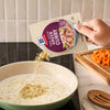 A hand pours a packet of Mc Thai Gn Curry Mix into a skillet of simmering coconut milk to prepare a quick meal.
