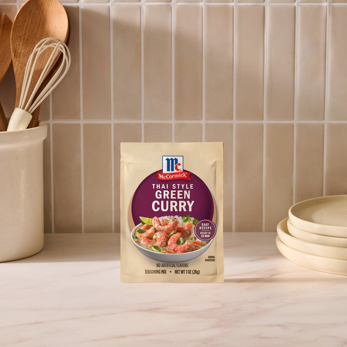 A package of Mc Thai Gn Curry Mix stands on a marble countertop in a kitchen setting with wooden utensils and stacked plates in the background.