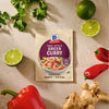 A package of Mc Thai Gn Curry Mix is centered on a textured surface surrounded by fresh ingredients like cilantro, limes, red peppers, and ginger root.