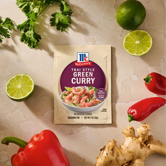 A package of Mc Thai Gn Curry Mix is centered on a textured surface surrounded by fresh ingredients like cilantro, limes, red peppers, and ginger root.