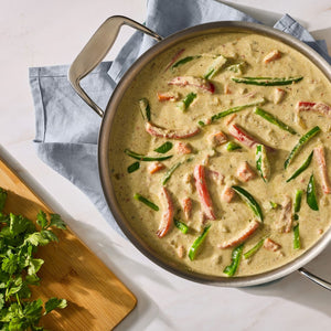 A large stainless steel pan filled with a creamy green curry featuring sliced red and green bell peppers prepared using Mc Thai Gn Curry Mix sits on a blue cloth next to fresh cilantro.
