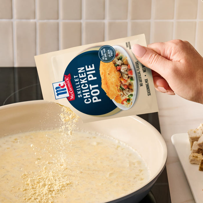A hand pours a packet of Mc Chkn Pot Pie Mix into a creamy sauce simmering in a skillet to create a quick one pan meal.
