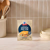 A package of Mc Chkn Pot Pie Mix stands on a marble countertop in a kitchen setting with wooden utensils and stacked plates.