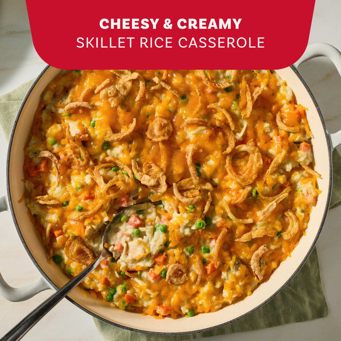 A large skillet filled with a cheesy and creamy rice casserole featuring peas and carrots topped with melted cheddar and crispy fried onions made using Mc Chkn Pot Pie Mix.
