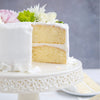 A two-layer vanilla cake with white frosting and floral decorations sits on a decorative white cake stand with a slice of cake on a plate in the background.