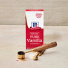 A box of McCormick Pure Vanilla Extract sits next to two gold measuring spoons one filled with vanilla extract.