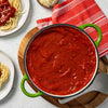 ['A pot of red sauce sits on a wooden cutting board with a red and white striped towel next to it.']