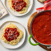 A pot of red sauce sits on a wooden cutting board with a red and white striped towel next to it.