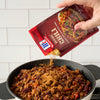 ['A hand pours McCormick Original Chili seasoning into a pot of chili.']