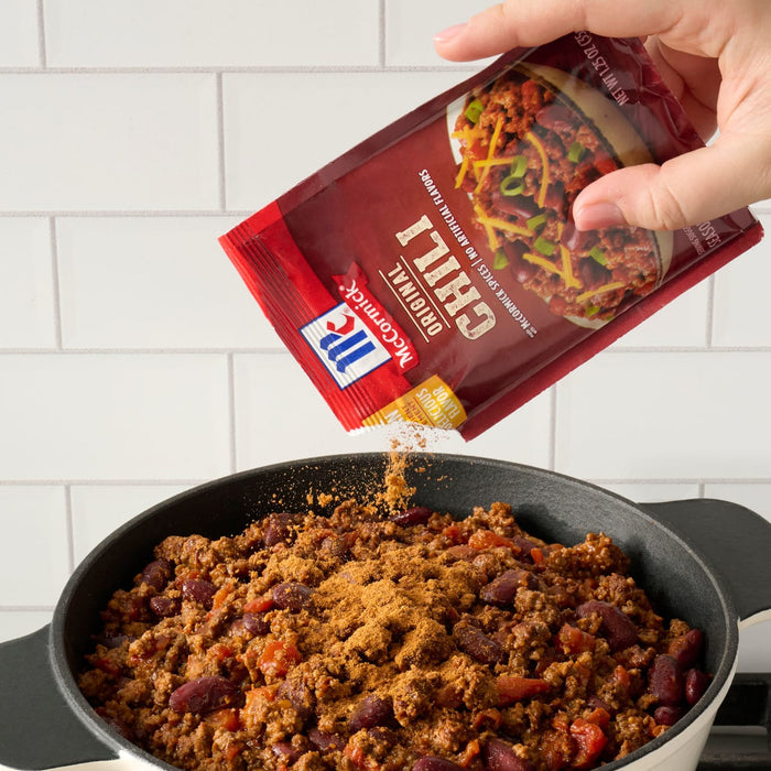 ['A hand pours McCormick Original Chili seasoning into a pot of chili.']