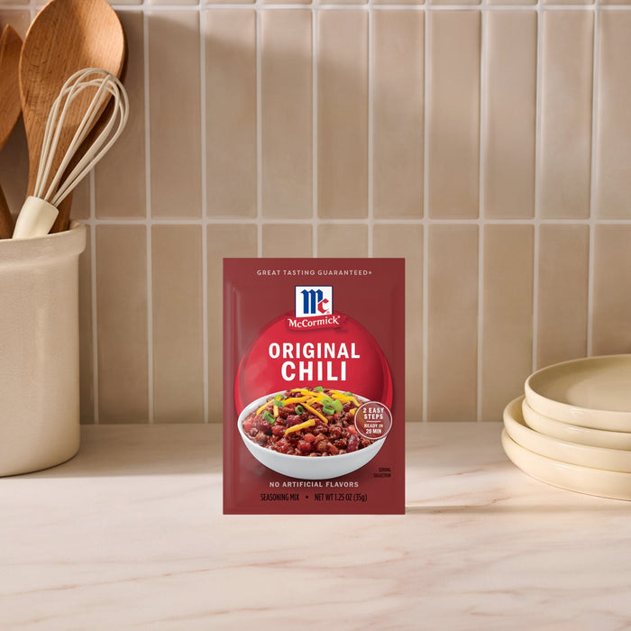 A package of Mccormick Chili Seasoning Mix 1.25 Oz stands upright on a white countertop, featuring a bowl of chili with cheese and green onions on its label, against a background of white subway tiles and kitchen utensils.