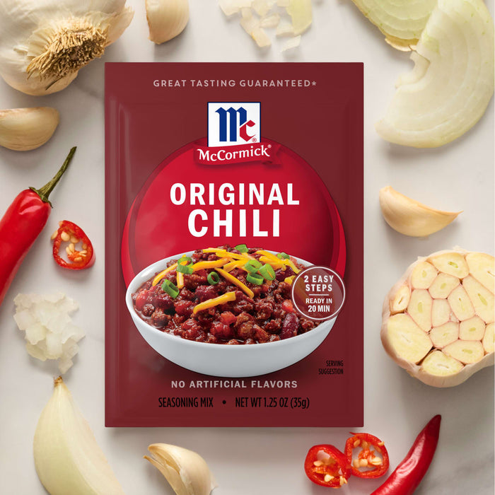 A Mccormick Chili Seasoning Mix 1.25 Oz packet is displayed on a white surface, surrounded by fresh ingredients like garlic, onions, and red chili peppers.