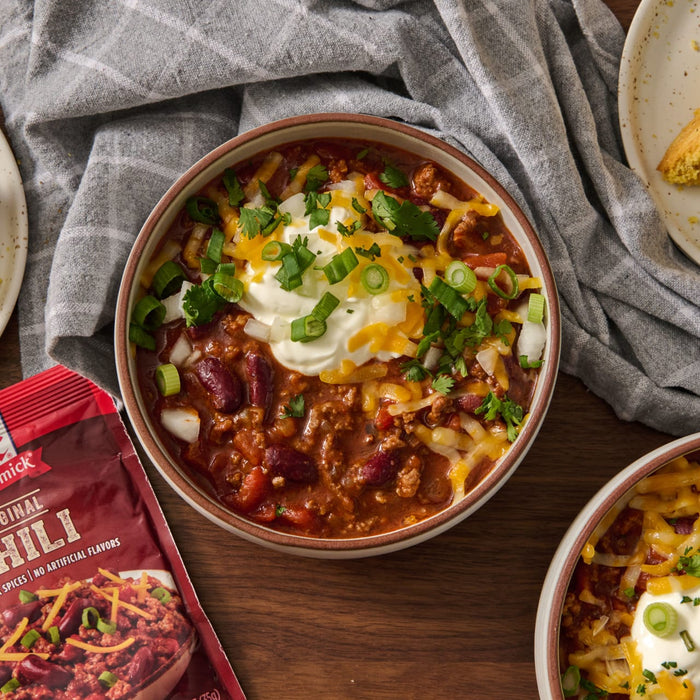 ['A bowl of chili topped with sour cream, shredded cheese, and green onions, with a McCormick Original Chili seasoning packet in the foreground.']