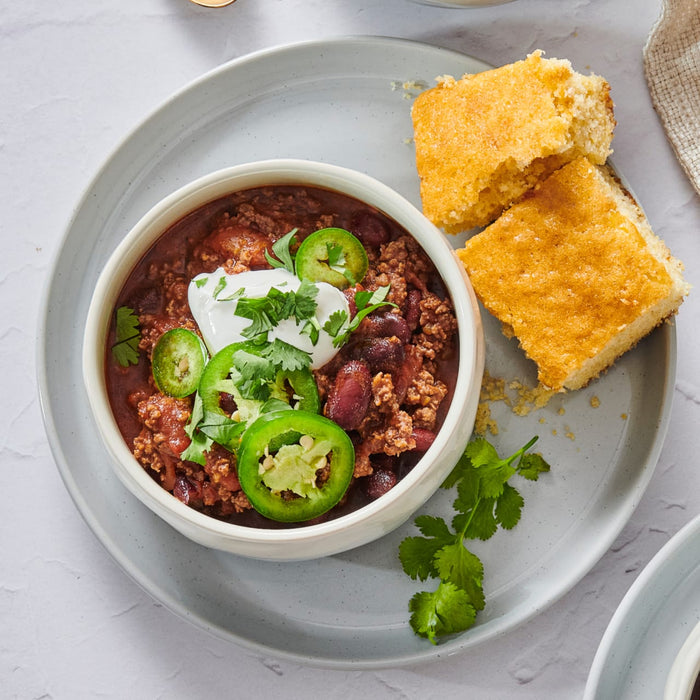 A hearty bowl of chili, garnished with shredded cheese, sour cream, and green onions, sits beside a package of Mccormick Chili Seasoning Mix 1.25 Oz on a wooden table with a gray cloth and cornbread.
