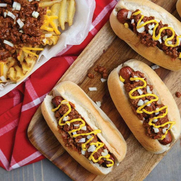 ['Three hot dogs topped with chili, onions, and mustard on a wooden cutting board.']