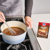 ["A person's hand stirs a pot of McCormick Au Jus Gravy on a stovetop."]