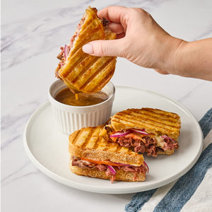 ['A hand dips a grilled cheese sandwich into a bowl of au jus.']