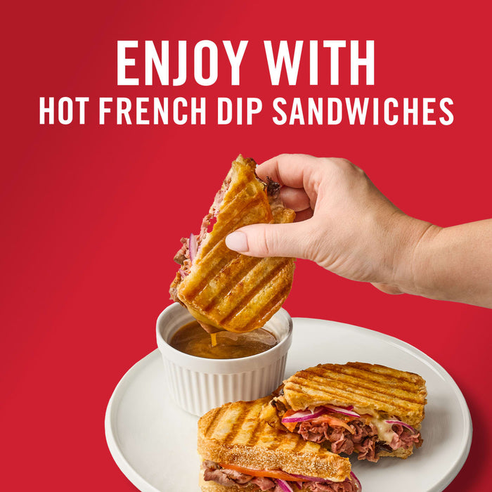 ['A hand dips a hot french dip sandwich into a bowl of au jus.']