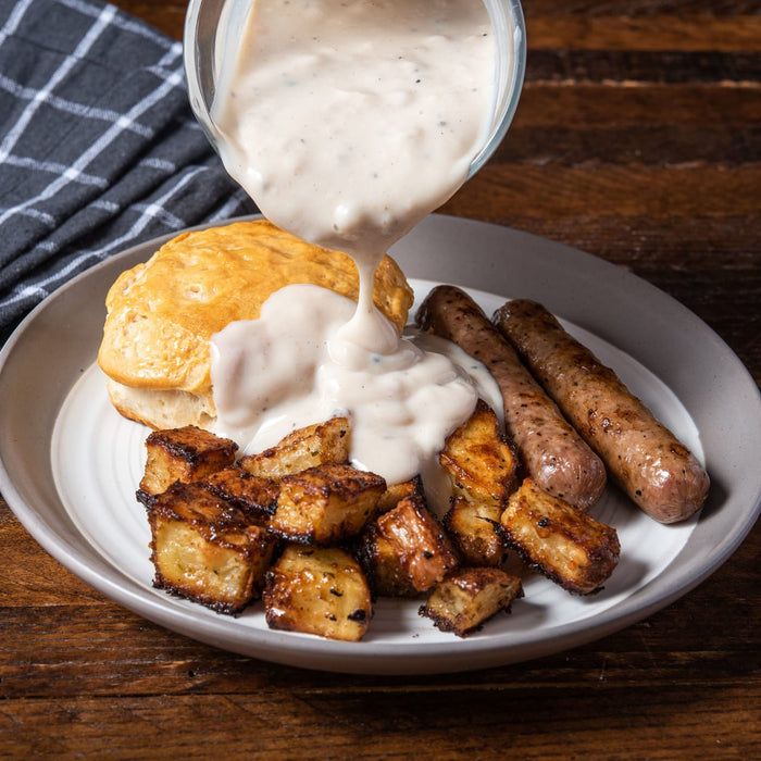A plate of sausage, potatoes, and biscuits with gravy being poured over it.