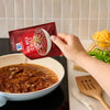 A person pours a packet of Mccormick Tex Mex Chili Seasoning Mix 1.25 Oz into a simmering pan of chili on a stovetop next to bowls of shredded cheese and sliced jalapeños.