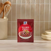 A packet of Mccormick Tex Mex Chili Seasoning Mix 1.25 Oz stands on a marble countertop in a kitchen setting with wooden utensils and ceramic bowls in the background.