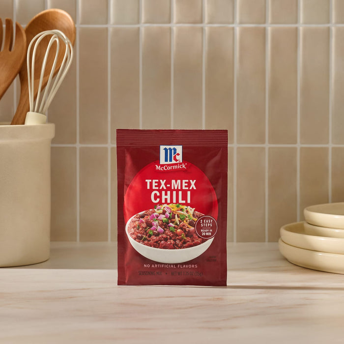 A packet of Mccormick Tex Mex Chili Seasoning Mix 1.25 Oz stands on a marble countertop in a kitchen setting with wooden utensils and ceramic bowls in the background.