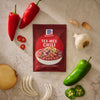 A package of Mccormick Tex Mex Chili Seasoning Mix 1.25 Oz is centered on a stone surface surrounded by fresh ingredients like red and green peppers, garlic cloves, and a whole onion.