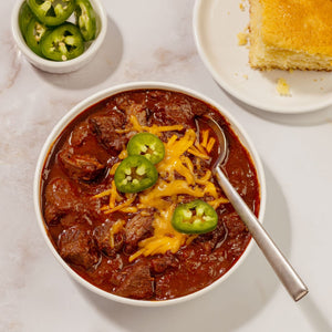 A hearty bowl of beef chili topped with melted cheddar cheese and sliced jalapeños is prepared using Mccormick Tex Mex Chili Seasoning Mix 1.25 Oz and served with a side of cornbread.