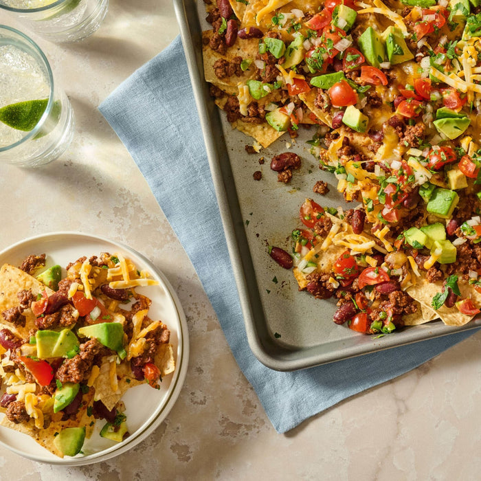 A sheet pan and a small plate are filled with loaded beef nachos featuring ingredients seasoned with Mccormick Tex Mex Chili Seasoning Mix 1.25 Oz and topped with avocado and tomatoes.