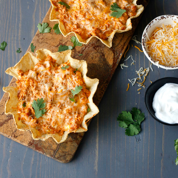['Two tortilla bowls filled with cheesy chicken dip, garnished with cilantro, and served with shredded cheese and sour cream.']