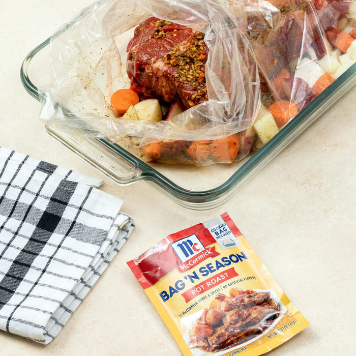 ["A package of McCormick Bag'N Season Pot Roast seasoning mix with a glass baking dish containing a raw pot roast and vegetables inside a cooking bag."]