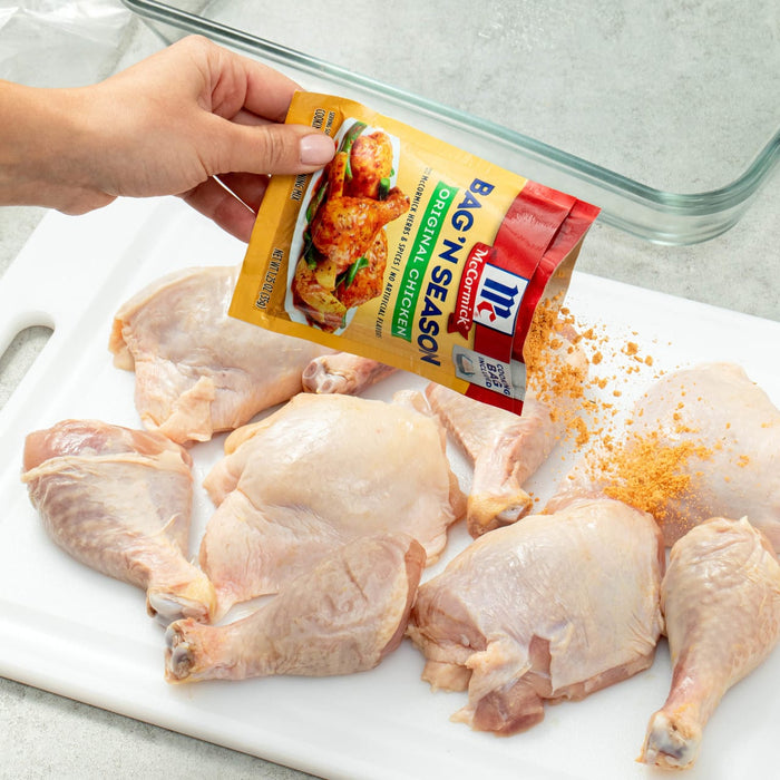 ["A hand is pouring McCormick Bag 'N Season Original Chicken seasoning onto raw chicken pieces."]