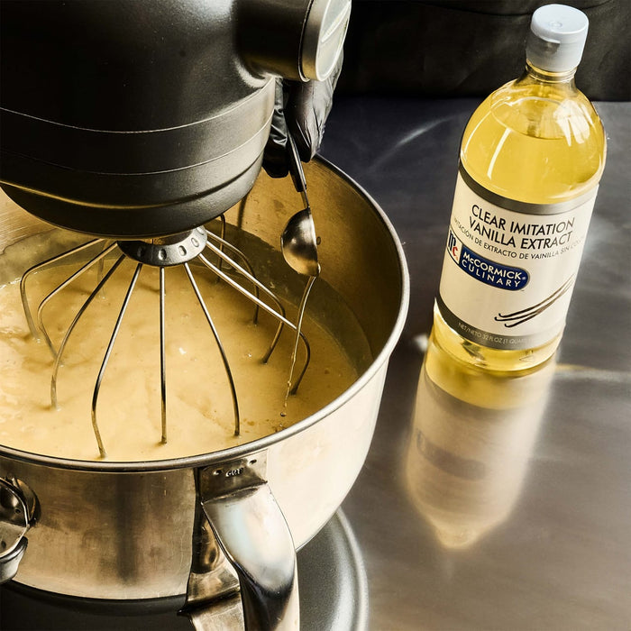 A close-up of a hand pouring McCormick Culinary Clear Imitation Vanilla Extract into a stand mixer.