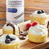 A bottle of McCormick Culinary Clear Imitation Vanilla Extract next to a wooden board with mini cheesecakes topped with whipped cream and various toppings.