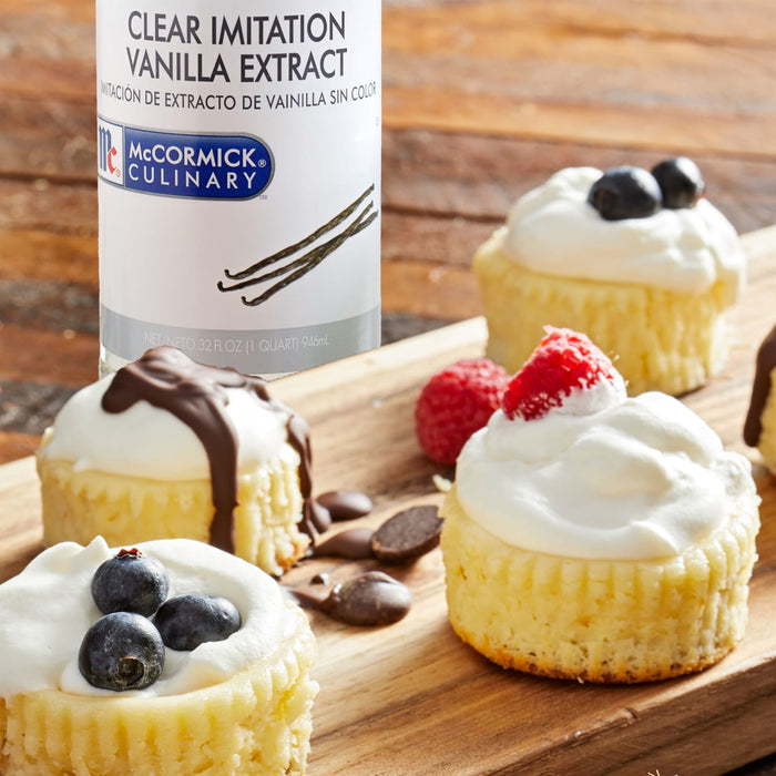 A bottle of McCormick Culinary Clear Imitation Vanilla Extract next to a wooden board with mini cheesecakes topped with whipped cream and various toppings.
