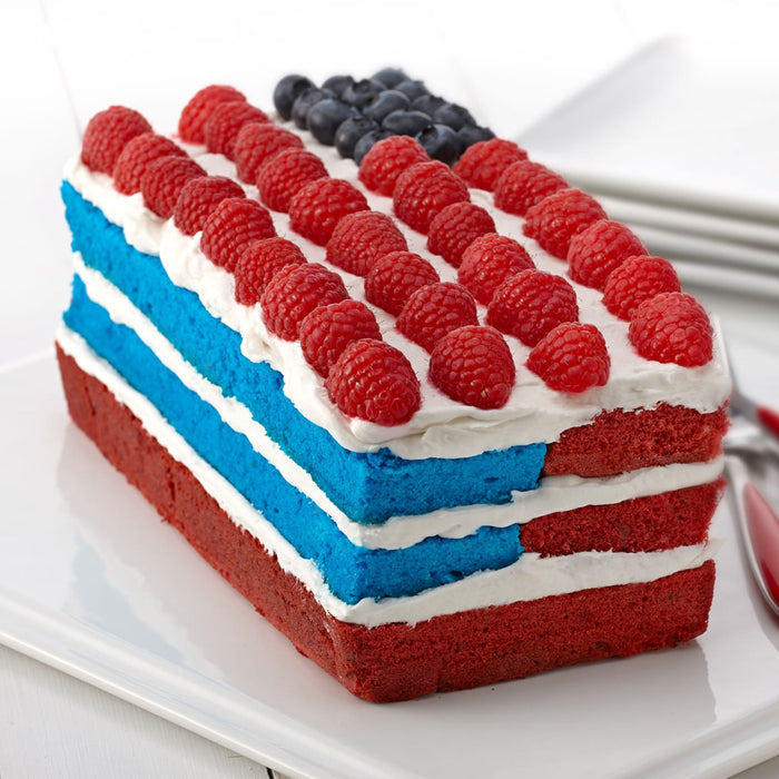 A vibrant layered cake, featuring blue layers likely colored with Mccormick Culinary Blue Food Coloring, and red layers, is decorated to resemble an American flag with white frosting, raspberry stripes, and a blueberry field.