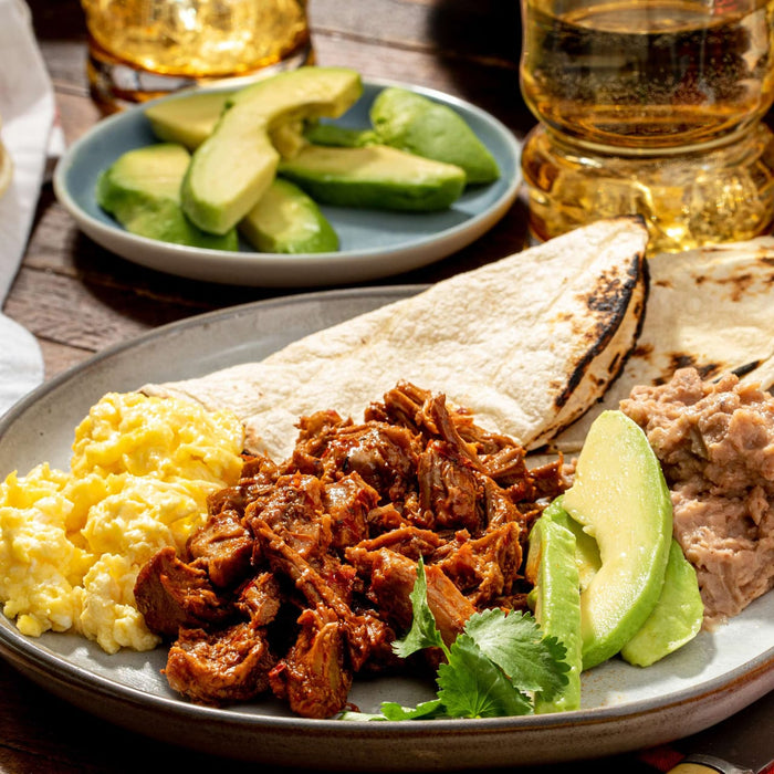 A plate of Mexican breakfast with scrambled eggs, chorizo, tortillas, refried beans, and avocado slices.