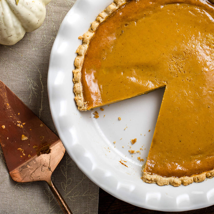 A pumpkin pie with a slice removed sits on a white plate next to a pie server.