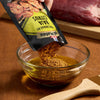 A hand pours McCormick Baja Citrus marinade mix into a bowl of oil, preparing a marinade for meat.
